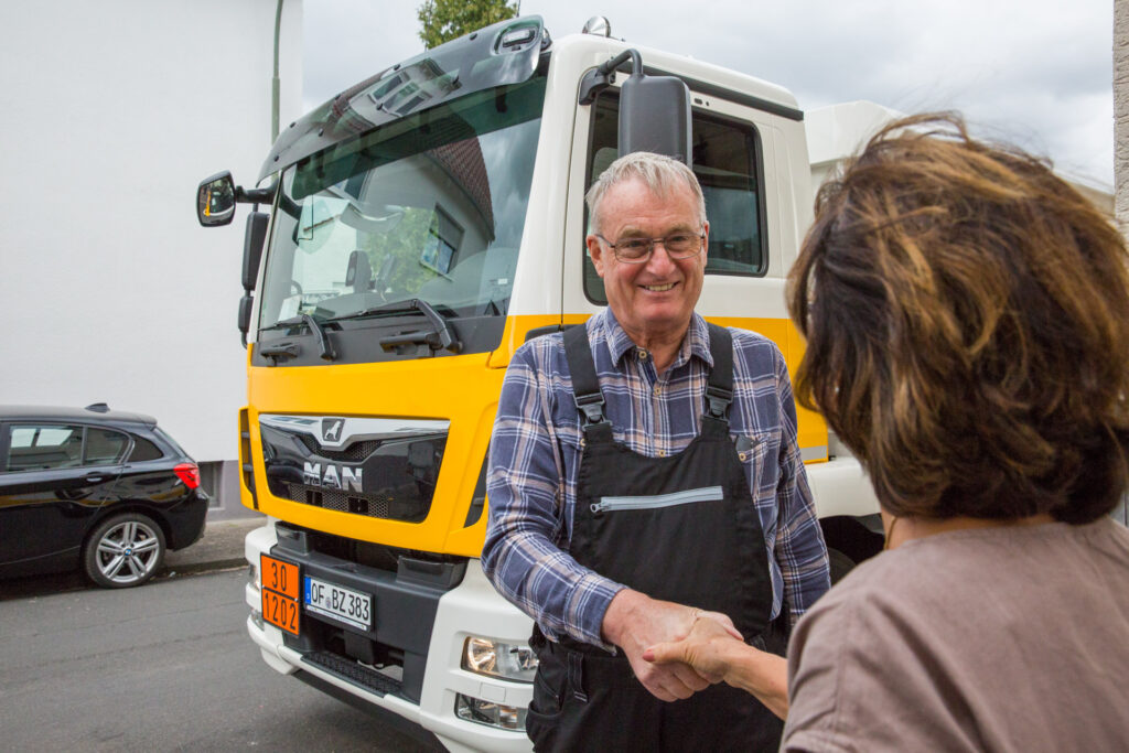Mann in Arbeitskleidung schüttelt einer Frau vor einem gelb-weißen Heizöl-Tankwagen die Hand